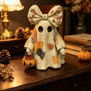 Könnte beinhalten: Eine cremefarbene Geisterfigur mit einer großen Schleife auf dem Kopf, die mit rosa Rosen verziert ist. Der Geist hat ein Patchwork-Muster in Orange-, Grau- und Brauntönen und hält einen kleinen orangefarbenen Kürbiseimer in einer Hand.