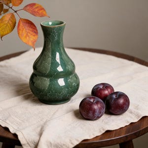 Könnte beinhalten: Eine grüne Keramikvase mit bauchiger Doppel-Kürbis-Form und glänzender, rissiger Glasur. Die Vase steht neben drei dunkelvioletten Pflaumen auf einem hellen Stofftuch, wobei im oberen Eck herbstliche Blätter zu sehen sind.