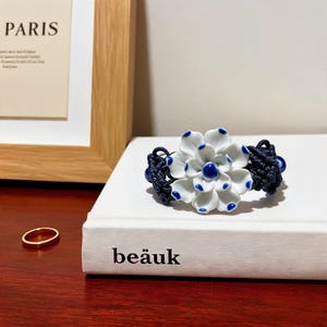 Könnte beinhalten: Ein Armband mit einer zentralen weißen Keramikblüte mit blau getippten Blütenblättern und einem blauen Zentrum, befestigt an einem dunkelblauen geflochtenen Kordelband. Das Kordelband enthält kleine blaue Kugeln im komplexen Knotendesign.