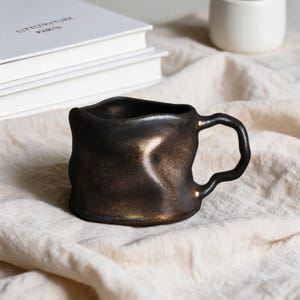 Op de afbeelding: Een zwarte keramische mok met een metallic bronzen afwerking en een onregelmatige, golvende, met de hand gevormde vorm. De mok heeft een gebogen handvat en een getextureerd oppervlak dat het licht opvangt en reflecties creëert op de donkere buitenkant.