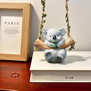 Könnte beinhalten: Eine graue Koala-Figur, die ein grünes Blatt hält und auf einer braunen Astschaukel sitzt, die an zwei mit kleinen grünen Blättern verzierten Seilen hängt. Der Koala hat eine schwarze Nase und ein strukturiertes Fell.