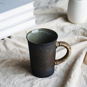 May include: A cylindrical ceramic mug with a dark, mottled black and brown exterior finish. The interior of the mug features a speckled light green glaze. A curved handle is wrapped in natural tan-coloured woven rope or twine material.