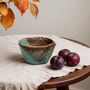May include: A small, hand-formed ceramic bowl featuring a textured exterior with a blend of turquoise and dark brown glaze. The bowl has an irregular, organic shape with a visible cross-hatched pattern on the side and a smooth, dark brown interior.