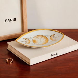 May include: An eye-shaped white ceramic trinket dish with a gold-rimmed edge. The centre features a gold-toned illustration of moon phases, including a full moon and crescent moons connected by dotted lines, resting on a white book with the text beauk.