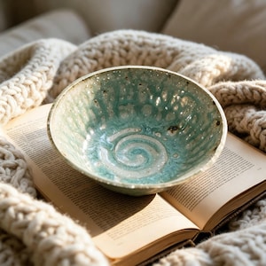 Könnte beinhalten: Eine runde Keramikschale mit einer texturierten, genoppten Oberfläche, die mit einer glänzenden meergrünen Glasur überzogen ist. Das Innere zeigt ein weißes Spiralmuster in der Mitte, einen gesprenkelten Rand und eine unebene Kante.