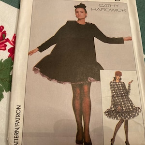 Può includere: Un modello di cucito vintage con due abiti per donne. Il primo abito è un abito nero con un bordo di pizzo bianco. Il secondo abito è un abito a quadri con un orlo a balze. Il modello è di Cathy Hardwick.