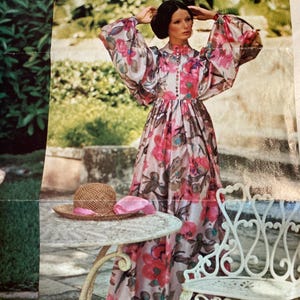 Peut inclure: Une femme portant une longue robe à fleurs rose et blanche avec une taille cintrée et des manches longues. Elle se tient dans un jardin avec une table et une chaise en arrière-plan.