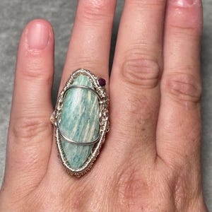 Puede incluir: Un anillo de alambre que presenta una gran piedra ovalada de color azul verdoso claro con vetas blancas. La piedra está envuelta en un intrincado trabajo de alambre plateado, con un pequeño acento de piedra preciosa redonda rosa oscuro.
