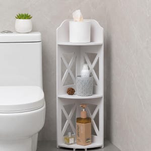 May include: A white three-tier corner storage shelf featuring X-shaped side panels. The top shelf holds a white tissue box, the middle shelf contains a grey textured cup and a small brown sphere, and the bottom shelf holds a soap dispenser and bottle.