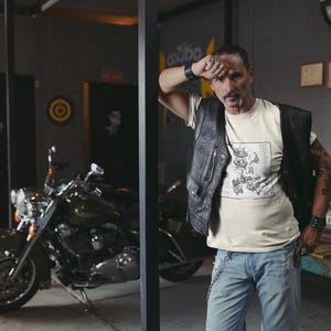 May include: A cream-colored short-sleeve t-shirt featuring a detailed black and white technical diagram of a motorcycle engine on the front, paired with a black leather vest, light blue denim jeans, and a silver chain attached to the waistband.