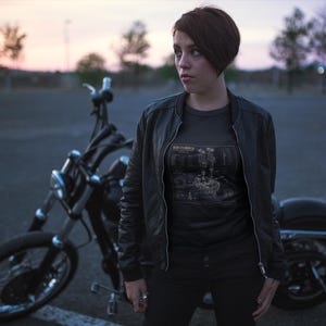 May include: A dark gray t-shirt featuring a detailed technical diagram of a motorcycle engine with the text "FOUR CYLINDER" printed on the front, worn under a black bomber jacket with a black motorcycle parked in the background.