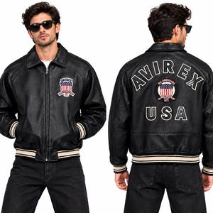 Puede incluir: Una chaqueta de cuero negra con cremallera frontal, cuello vuelto y puños y dobladillo de canalé a rayas. El pecho muestra un parche de bandera Avirex USA. La espalda presenta letras blancas grandes de Avirex, un parche circular y USA.