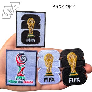 May include: A pack of 4 embroidered patches featuring FIFA World Cup 2026 designs. The set includes rectangular and shaped patches in light blue, black, and white, displaying the gold trophy, the text FIFA, and the text MEXICO USA CANADA.