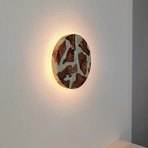 Op de afbeelding: Een ronde wandlamp met een mozaïek van bruine stukken hout ingebed in een doorschijnend, gebroken wit harsmateriaal. De lamp verspreidt een warme, zachte gloed vanaf de randen, wat een halo-effect op de muur creëert.