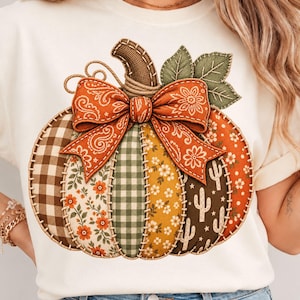 Può includere: Una t-shirt a maniche corte color crema con una grande grafica di una zucca composta da vari inserti simili a tessuto, tra cui quadretti marroni, stampe floreali, tartan verde e motivi a cactus, sormontata da un grande fiocco arancione.