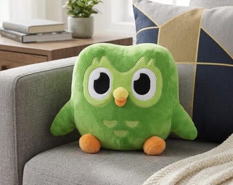May include: A plush toy shaped like a green owl with large white eyes, black pupils, and a small yellow beak. The owl has two orange feet and light green feather details on its chest. The soft, rounded figure features a vibrant green body.