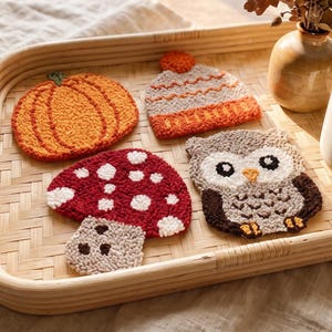 May include: A set of four punch needle coasters featuring an orange pumpkin, a grey and orange knit-style hat, a red and white mushroom, and a brown and white owl, arranged on a rectangular woven bamboo tray with a ceramic mug and vase.