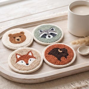 Pode incluir: Quatro porta-copos circulares com rostos de animais bordados, incluindo um urso castanho, um guaxinim cinzento, uma raposa laranja e um bisão castanho escuro. Os porta-copos estão dispostos numa bandeja de madeira ao lado de uma caneca.