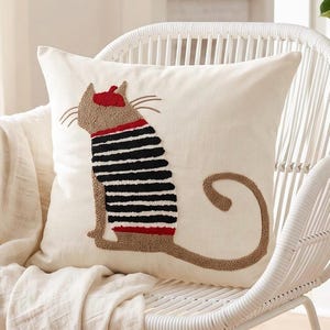 Op de afbeelding: Een vierkant gebroken wit sierkussen met een getextureerd punch needle ontwerp van een kat die een rode baret en een zwart-wit gestreept shirt draagt. De kat is in profiel afgebeeld met een lange gekrulde staart in beige, zwart en rood garen.