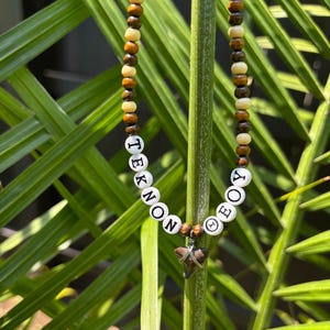 May include: A beaded necklace featuring alternating dark brown and light tan wooden beads. The necklace includes white circular beads with black lettering spelling out TEKNON and OEOY, centered with a small, dark, triangular shark tooth pendant.