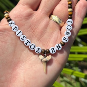 Op de afbeelding: Een kralenketting met afwisselend bruine houten kralen en witte ronde kralen met zwarte Griekse letters die XPIΣTOΣ EN EMOI spellen. Een kleine, lichtgekleurde haaientandhanger is met draad omwikkeld en hangt in het midden van de ketting.