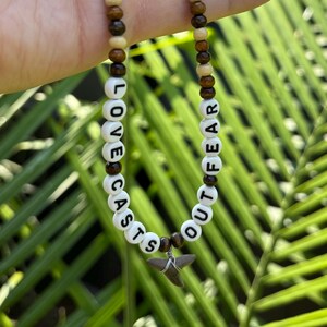 May include: A beaded necklace featuring alternating dark brown and light tan wooden beads. White letter beads spell out the phrase LOVE CASTS OUT FEAR. A small, grey, triangular shark tooth charm is wire-wrapped and suspended at the bottom center.