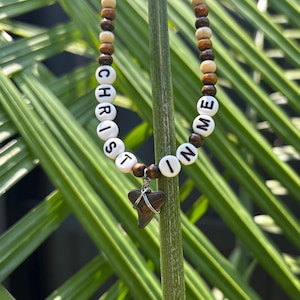 Op de afbeelding: Een kralenketting met afwisselend licht- en donkerbruine houten kralen en witte letterkralen die CHRIST IN ME spellen. Een donkerbruine haaientandhanger is omwikkeld met zilverdraad en hangt in het midden van de ketting.