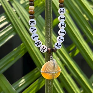 Op de afbeelding: Een ketting met een centrale gele schelphanger omwikkeld met zilverdraad, geflankeerd door twee strengen houten kralen in bruine en crèmetinten. De kralen bevatten witte ronde tussenstukjes met zwarte letters die KAINH en KTIΣIΣ spellen.