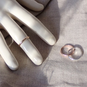 May include: A gold-colored ring is worn on a finger of a white hand. Two additional rings, one rose gold and one silver, are next to each other on a gray fabric surface. The rings are simple bands, showcasing minimalist jewelry.