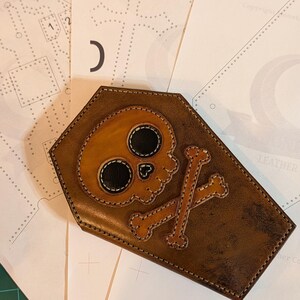 May include: A brown leather coffin-shaped case featuring a tan skull and crossbones appliqué with black eye sockets. The item is stitched with light-colored thread and rests on top of several white paper templates with printed patterns and markings.