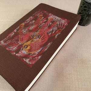 Peut inclure: Un livre à couverture rigide marron avec une impression graphique rectangulaire sur la couverture. L'image représente une main stylisée et abstraite dans des tons de rouge, orange et jaune sur un fond texturé sombre. Pages blanches.