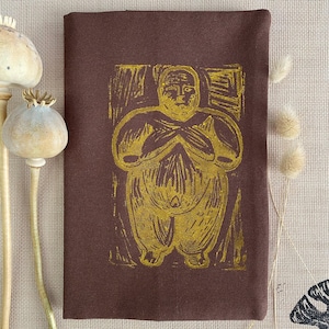 Pode incluir: Uma peça de tecido retangular castanho-escuro com uma ilustração central em bloco amarelo-mostarda de uma figura humana estilizada com os braços cruzados sobre o peito. A figura tem um tronco e pernas arredondados, representados com linhas gravadas.