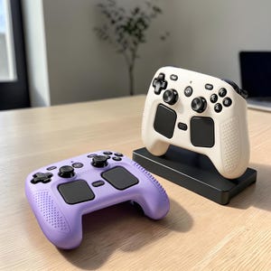May include: Two wireless video game controllers, one in cream and one in light purple, featuring black joysticks, directional pads, action buttons, and large touch-sensitive trackpads. The cream controller rests on a black rectangular charging dock.