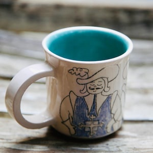 Peut inclure: Une tasse en céramique avec un extérieur crème brillant et un intérieur turquoise vif. La surface extérieure présente une illustration à l'encre noire d'un personnage historique portant un chapeau à larges bords et un gilet bleu avec une croix.