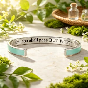 May include: A silver-toned cuff bracelet featuring a smooth, light blue enamel inlay along the outer band. The interior surface of the metal bracelet is engraved with the black text, "this too shall pass BUT WTF?!" in a clear, sans-serif font.