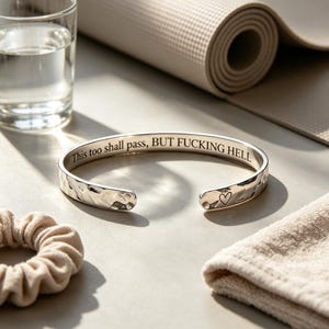 Puede incluir: Un brazalete de metal plateado con una textura martillada en el exterior. El interior está grabado con el texto "This too shall pass, BUT FUCKING HELL" y un pequeño símbolo de corazón está grabado cerca de uno de los extremos abiertos.