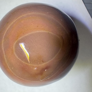 May include: A smooth, spherical object with a polished surface in shades of muted brown and tan. The sphere features subtle, concentric circular banding patterns across its surface, with a reflective highlight visible near the center.