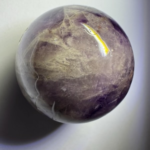 May include: A polished spherical stone featuring a mix of deep purple and translucent white mineral inclusions. The surface displays natural veining and internal fractures, with a bright white light reflection visible on the upper right side.