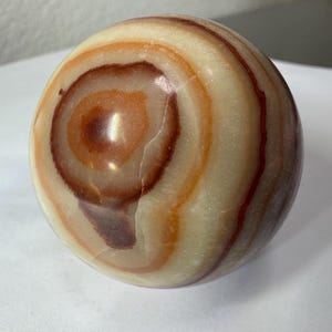 May include: A polished stone sphere featuring concentric bands of cream, tan, and deep reddish-brown colours. The smooth, rounded surface displays natural mineral layering with a glossy finish, creating a distinct circular pattern across the object.