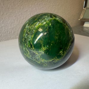 May include: A polished spherical stone with a deep forest green base color, featuring intricate, swirling patterns of lighter lime green and yellow mineral inclusions across its smooth, reflective surface, creating a marbled appearance.