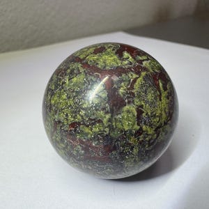 May include: A polished stone sphere featuring a mottled pattern of vibrant lime green and deep maroon red. The surface of the orb displays a complex, interconnected network of dark veins and crystalline textures, creating a smooth, reflective finish.