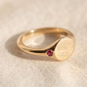 May include: A gold signet ring featuring a smooth, circular flat top and a single small, round, deep pink gemstone set into the band just below the main circular face. The metal has a polished, reflective finish with warm yellow tones.
