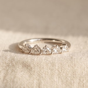 Op de afbeelding: Een zilverkleurige ring met vijf in klauwen gezette, driehoekig geslepen heldere stenen die in een rij op de bovenste helft van de band zijn geplaatst. Het metaal heeft een gepolijste, reflecterende afwerking en een geometrisch ontwerp.