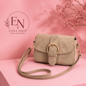 May include: A beige suede crossbody bag featuring a gold-tone buckle closure on the front flap and a matching adjustable shoulder strap. The bag is displayed with the logo EN ENNA SHOP BAGS & ACCESSORIES visible in the upper left corner.