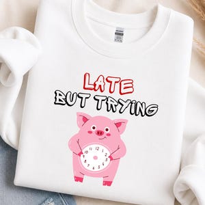 May include: A white crewneck sweatshirt featuring a graphic of a pink pig holding a clock. Above the pig, the text reads LATE in red and BUT TRYING in black. The sweatshirt has a standard ribbed collar and long sleeves, with a Gildan brand label visible.