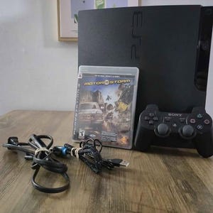 May include: A black PlayStation 3 console with a matching black Sony wireless controller, a MotorStorm game case, and two black power and charging cables. The console features the PS3 logo on the front panel and the Sony logo on the controller.