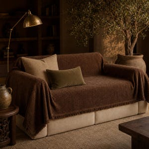 May include: A sofa covered with a dark brown textured throw blanket featuring fringe edges. The sofa is accented with one large square beige pillow and one smaller rectangular olive green velvet pillow, creating a layered and cozy seating arrangement.
