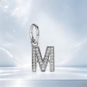 May include: A silver-toned letter M charm pendant featuring a polished metal finish and a front surface encrusted with numerous small, clear, round-cut stones. The charm includes a circular jump ring attached to the top for easy attachment.