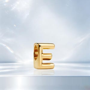 May include: A polished gold-tone metal charm in the shape of the uppercase letter E. The charm features a smooth, rounded, tubular design with a hollow center opening on the left side, designed to slide onto a bracelet or necklace chain.