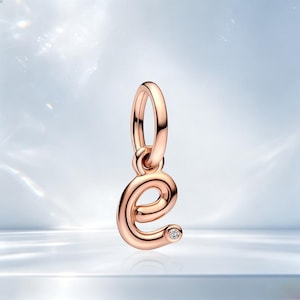 May include: A rose gold charm in the shape of the lowercase letter e, featuring a smooth, polished metallic finish and a single small, clear round gemstone set at the end of the letter's curve, suspended from a matching rose gold jump ring.